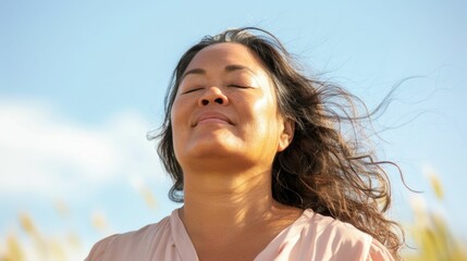 A woman with closed eyes smiling and her hair blowing in the wind against a blue sky.