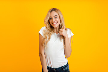 A young woman with long, blonde hair smiles and gives a thumbs up while standing in front of a bright yellow background. She is wearing a white t-shirt and blue jeans.