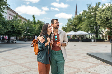 Fototapeta premium Young happy hipster backpacker travelers couple, visiting new country, summer travel destination, enjoying their vacation, walking down the city streets, looking historical buildings and architecture.