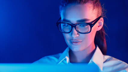 A woman wearing glasses is sitting indoors at night, focused on her laptop screen. The room is...