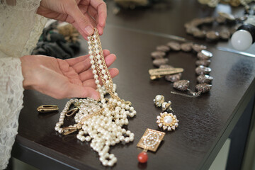 A person holds a pearl necklace over a table filled with various pieces of jewelry. This image...