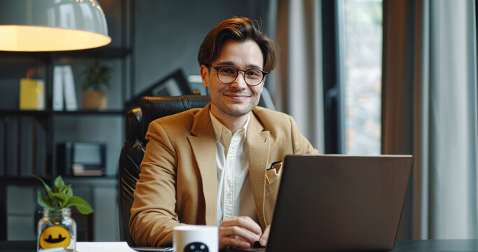 A man in a tan jacket is sitting at a desk with a laptop and a mug