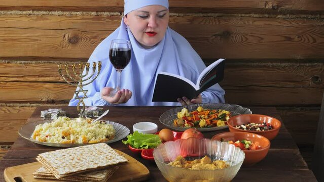 A Jewish woman in a blue veil with glasses of wine and siddur reads kiddush for wine on the Pesach Seder holiday at a set table. Medium plan