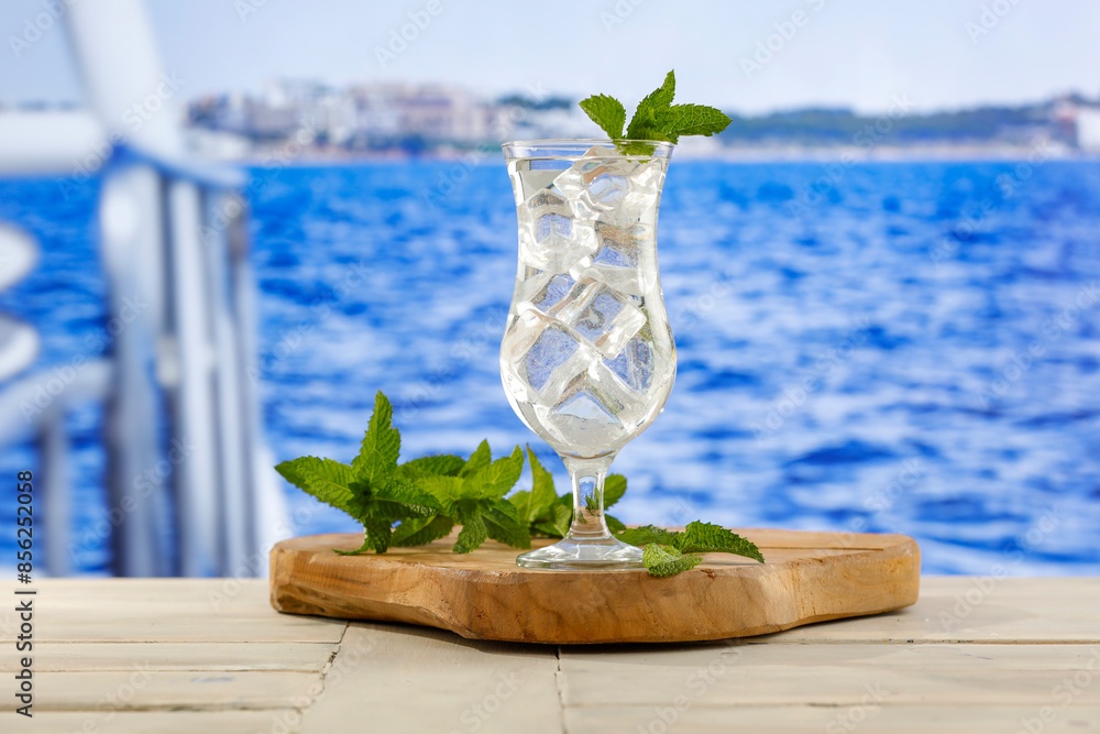 Wall mural glass of cold icy mineral water on wooden table top with beautiful landscape of sea and blue sky bac