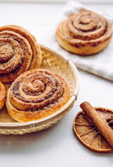 Freshly baked homemade cinnamon rolls in basket in home kitchen on white table, cinnamon stick for decoration. Selective focus, side view, natural back light.