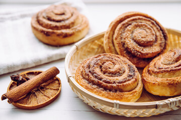 Freshly baked homemade cinnamon rolls in basket in home kitchen on white table, cinnamon stick for decoration. Selective focus, side view, natural back light.
