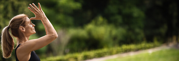 Side view of sporty young blonde woman practicing meditation at public park, holding hands next to face, eyes closed, enjoying yoga workout outdoors, closeup, panorama with copy space