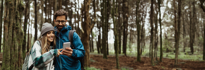 A young couple is hiking through a forest and looking at a smartphone. The man is wearing glasses and a blue jacket, and the woman is wearing a grey beanie and a plaid jacket, copy space