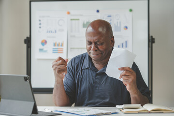 A senior man at a desk looking stressed while reviewing office documents with a laptop. Workplace stress, analysis, and business reports concept.