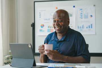 Smiling man drinking coffee and working on laptop at home office. Charts and graphs displayed on board in background. Remote work concept.