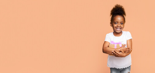 A little girl with a big smile holds a wrapped gift box in front of a peach-colored background. She is wearing a white t-shirt and denim shorts. The girl has dark skin and short, curly brown hair.