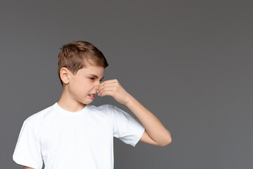 A young boy wearing a white t-shirt is pinching his nose, an expression of disgust on his face as...