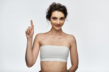 A young woman in a white strapless top points upwards with a confident smile.