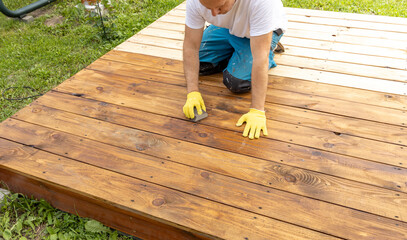 A man is kneeling on a wooden deck and staining it a rich brown color. He is wearing yellow gloves...