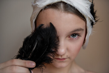 Young ballerina girl with feathered headband tiara holds a black feather over her face, eyes...