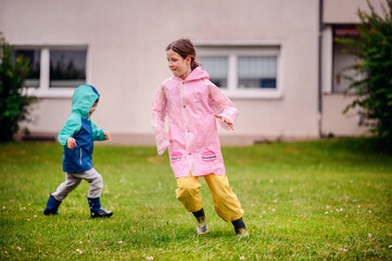 Two children, one in a teal and blue raincoat and the other in a pink raincoat, playing joyfully on a grassy field on a rainy day. The scene captures the fun and excitement of childhood.