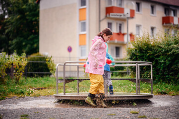 Two children in raincoats, one in pink and one in teal and blue, playing at a playground on a rainy day. The joyful scene captures them jumping and exploring with excitement.