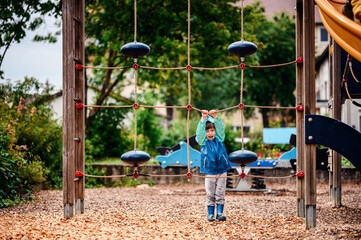 Young child in a teal and blue raincoat, hanging on a rope net at a playground on a rainy day. The scene captures their adventurous and playful spirit.