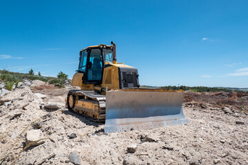 Perspectiva lateral de um bulldozer em operação num terreno pedregoso sob um céu azul © LuIvDa