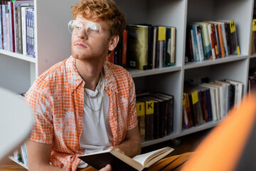 A young man engrossed in reading a book while seated in front of a bookshelf.