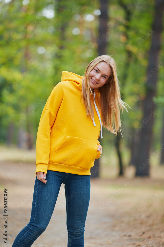 Wall mural Blue-eyed blonde in a yellow hoodie walks in a pine forest. Portrait of a joyful young woman enjoying in autumn park. - Wall murals