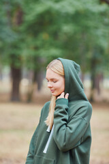 Blue-eyed blonde in a green hoodie walks in a pine forest. Portrait of a joyful young woman enjoying in autumn park.