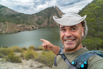 Adventurous hiker smiles looking at the camera, takes a selfie with a beautiful mountainous landscape with a lake in the background.