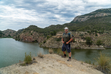  A strong man hiker poses smiling on a rock, with a landscape with mountains and a lake in the background