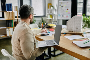 A bearded businessman works on a laptop at a cluttered desk in a modern office.