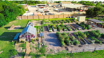 Urban community garden with glass greenhouse near large manufacturing in North Dallas, row of wooden metal raised beds, storage sheds, patch adopt a plot group gardening food bank, Texas, aerial