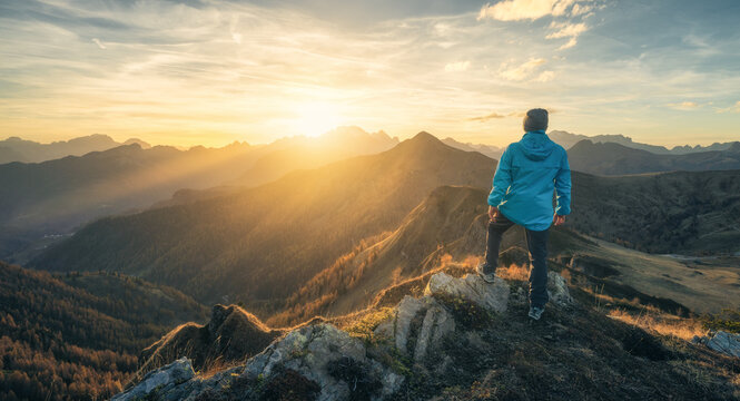 Man on stone on the hill and beautiful mountain valley in haze at colorful sunset in autumn. Dolomites, Italy. Guy, mountain ridges in fog, orange grass and trees, blue sky with sun in fall. Hiking