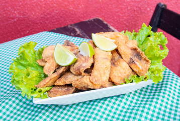 Portion of crunchy breaded deep fried fish in cubes, with lettuce below and lemon on top, on a table, in close-up.