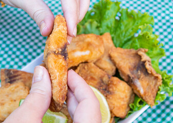 Hands holding piece of crunchy breaded deep fried fish on top of portion of crunchy breaded deep fried fish in cubes, with lettuce below and lemon on top, on a table, in close-up.