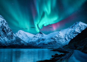 Northern lights over the snowy mountains, frozen sea, reflection in water at winter night in Lofoten, Norway. Aurora borealis and snowy rocks. Landscape with polar lights, road, starry sky and fjord