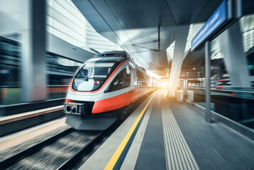 High-speed orange passenger train moving at railway station platform at sunset. Train station....