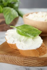 Piece of bread with cream cheese and basil leaf on white table, closeup