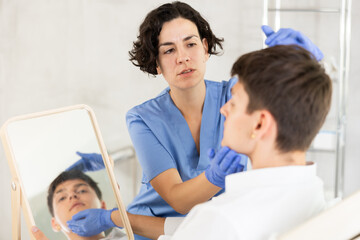 Young man sits in chair at a doctor appointment with beautician. Woman doing facial examination before surgery