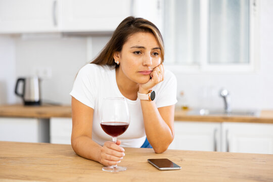Sad frustrated young hispanic woman drinking wine and waiting for call while standing alone in home kitchen, moile phone lying on table. Loneliness concept