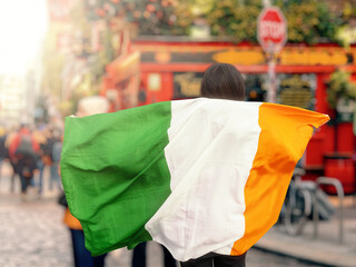 Teenager girl holding National flag of Ireland in her extended hands, busy street and pub in the...
