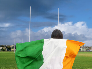 Teenager girl standing and holding National flag of Ireland. Selective focus. Tall goal posts and...