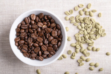 Roasted coffee beans in a white bowl on linen fabric. On the right unroasted green Arabica coffee beans, seeds of berries from Coffea arabica, also Arabian, mountain or arabica coffee. Close-up. Photo