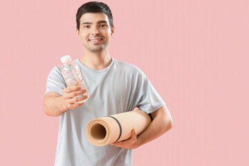 Sporty young man with bottle of water and fitness mat on pink background