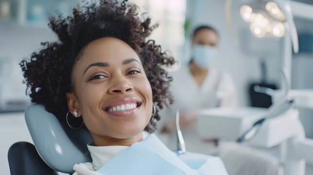 Cheerful black woman with curly hair smiling in dental chair, with focus on her while dentist in background, bright clinic setting.