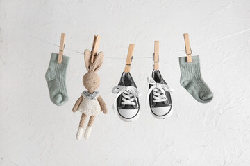 Stylish baby shoes with cute toy and socks hanging on rope against grunge white background