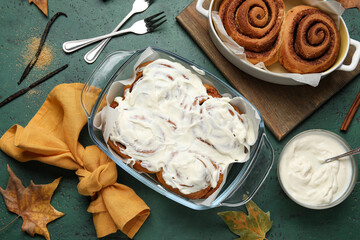 Baking dishes of tasty cinnamon rolls with cream and autumn leaves on green background