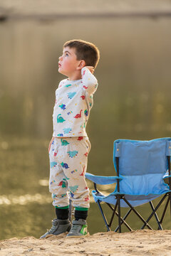 A young boy in his pyjamas standing on a river bank next to a folding chair looking up into the sky