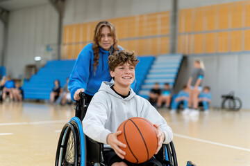 Naklejka premium A young man in a wheelchair dribbles a basketball while smiling and looking at the camera. A young woman stands behind him, pushing his wheelchair
