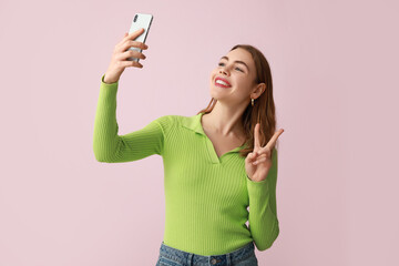 Pretty young woman showing peace gesture while taking selfie on pink background