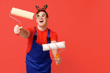 Portrait of cheerful male painter in Christmas reindeer headband with rollers on red background