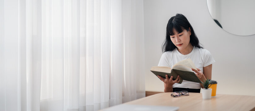 A woman is reading a book at a table. The book is open to a page with a picture of a woman on it. The woman is wearing a white shirt and is sitting in front of a window. There is a cup